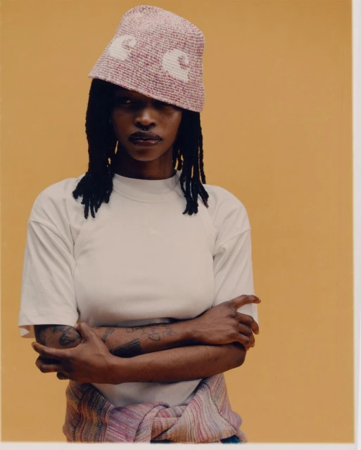 A young adult Black woman wearing a bucket hat poses against a mustard-yellow seamless backdrop in a medium studio portrait.