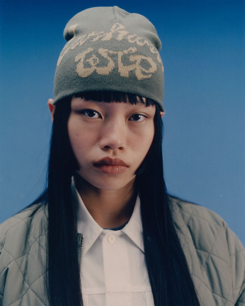 Portrait of an East Asian woman wearing a sage green knit beanie and matching quilted jacket against a light blue backdrop.