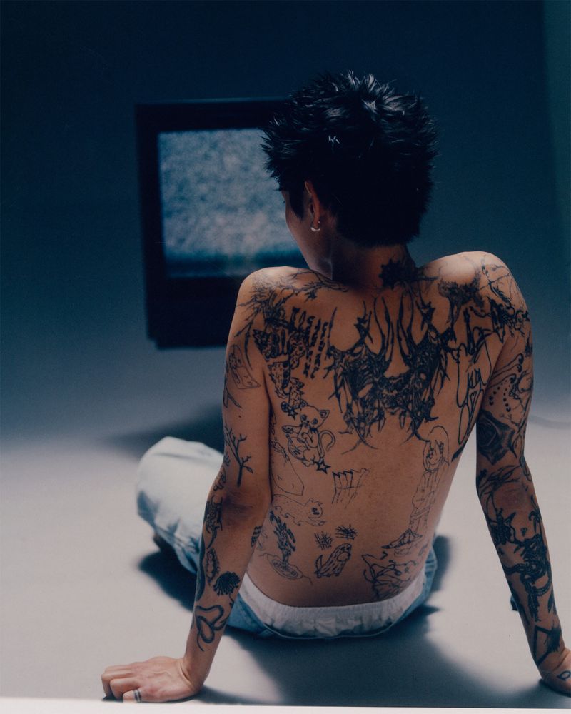 An adult human with tattoos sits on a floor and faces a static-filled television, indoors, with a medium camera shot in soft lighting.
