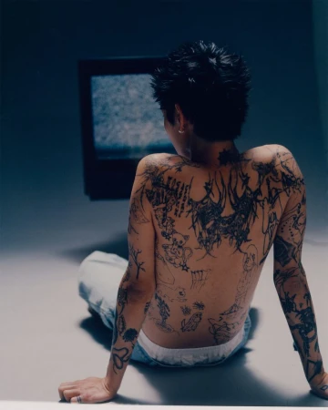An adult human with tattoos sits on a floor and faces a static-filled television, indoors, with a medium camera shot in soft lighting.