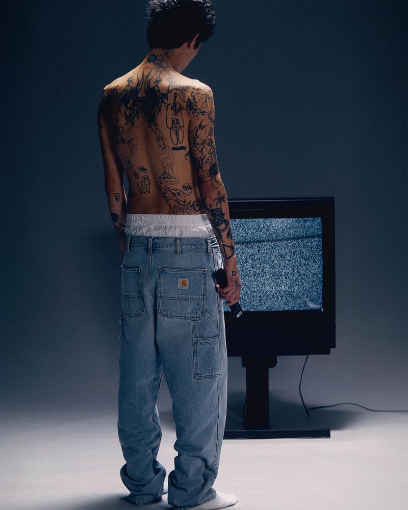 Tattooed man in loose Carhartt jeans stands in front of static TV screen in a softly lit studio, holding a black remote.