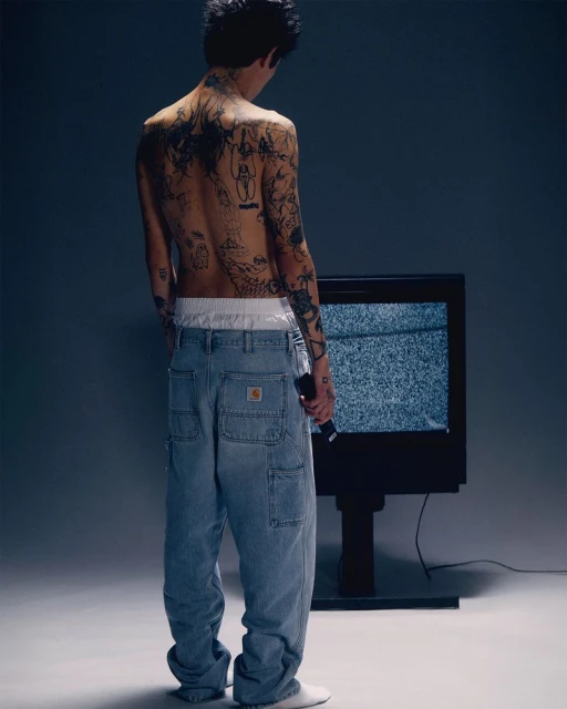 Tattooed man in loose Carhartt jeans stands in front of static TV screen in a softly lit studio, holding a black remote.