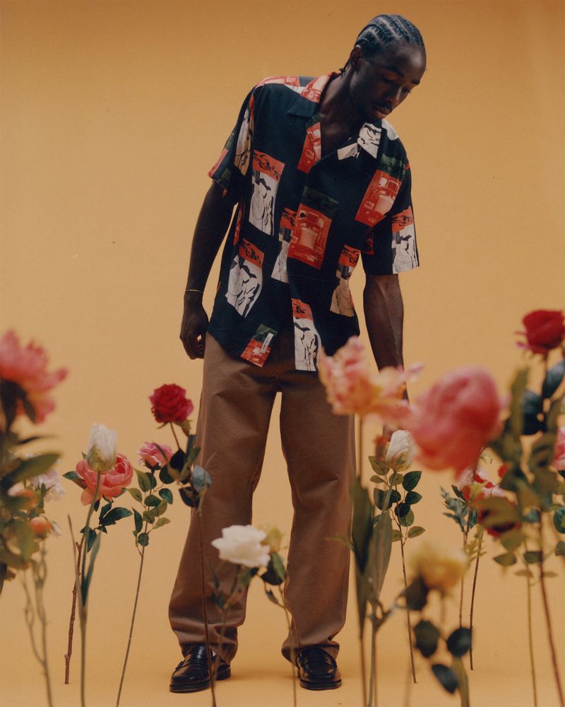 Man in patterned shirt and tan pants stands in field of flowers against yellow backdrop in fashion shot, looking down, low angle.
