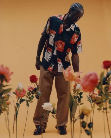 Man in patterned shirt and tan pants stands in field of flowers against yellow backdrop in fashion shot, looking down, low angle.