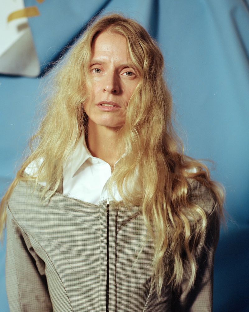 Blonde woman in white shirt and grey jacket poses against light blue backdrop in portrait shot, captured in studio lighting.