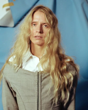 Blonde woman in white shirt and grey jacket poses against light blue backdrop in portrait shot, captured in studio lighting.