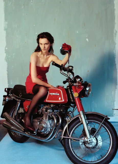 Adult woman in red dress poses on red Honda motorcycle against mottled blue and white background in a studio setting. Automotive fashion.
