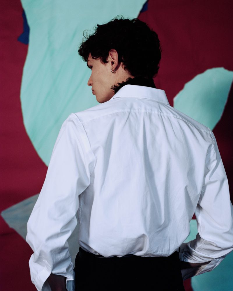 Young man seen from behind wearing a crisp white button-down shirt against a backdrop with maroon and light blue abstract shapes.