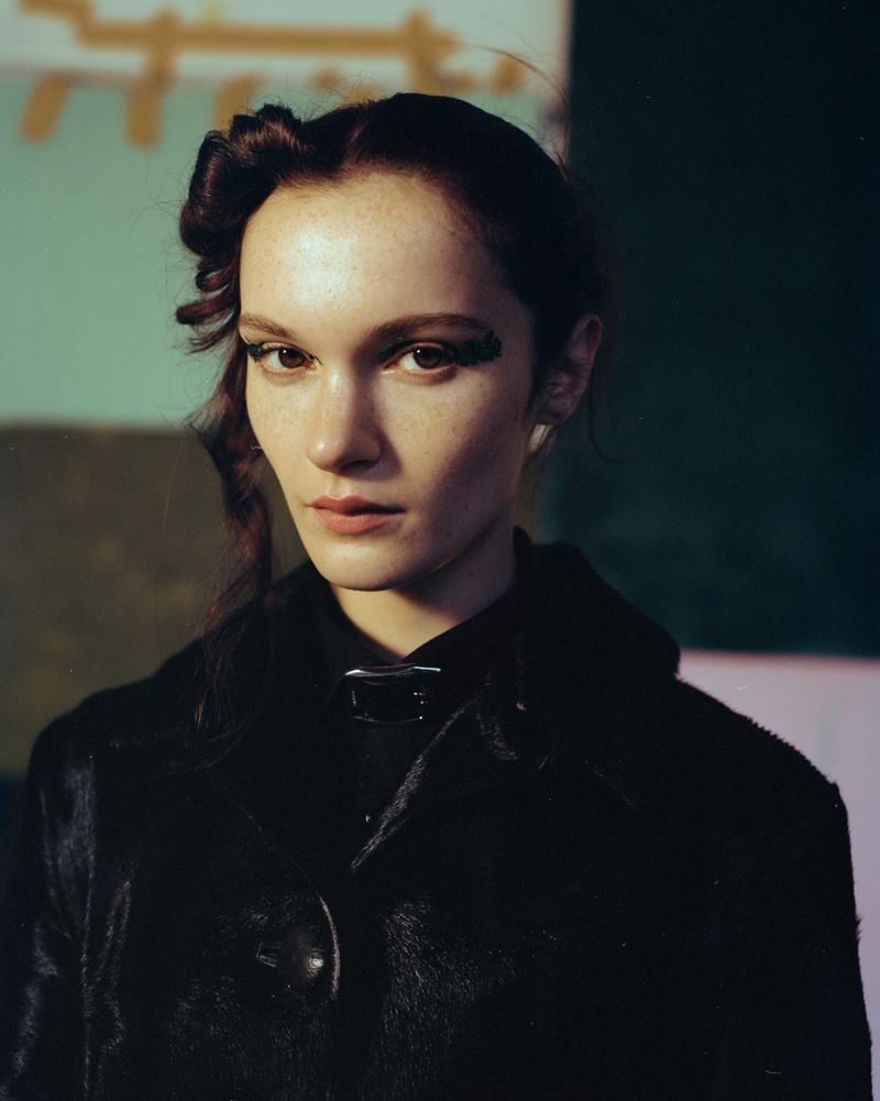 Adult woman in a black fur coat and dramatic eye makeup poses indoors, close-up shot, with a painted backdrop in muted tones.