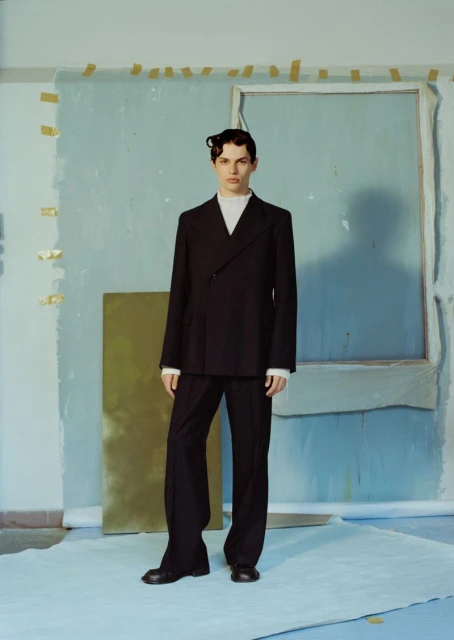 Fashion portrait of a young adult male posing in a double-breasted black suit with a white shirt in a studio setting against a blue backdrop.