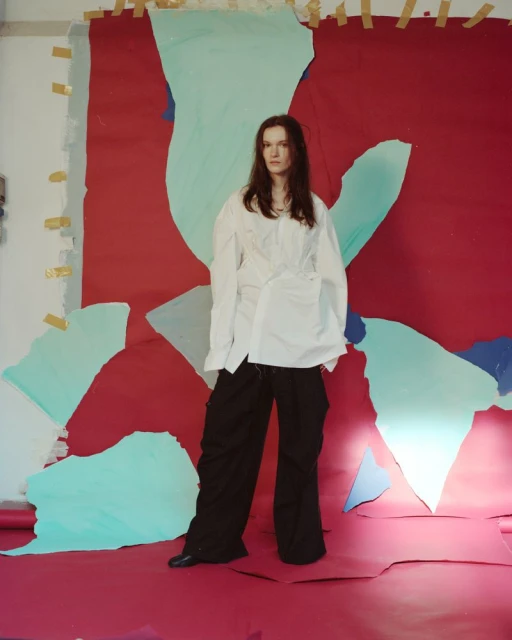 Woman in oversized white shirt and black pants stands in front of a colorful abstract painted backdrop in a studio setting.