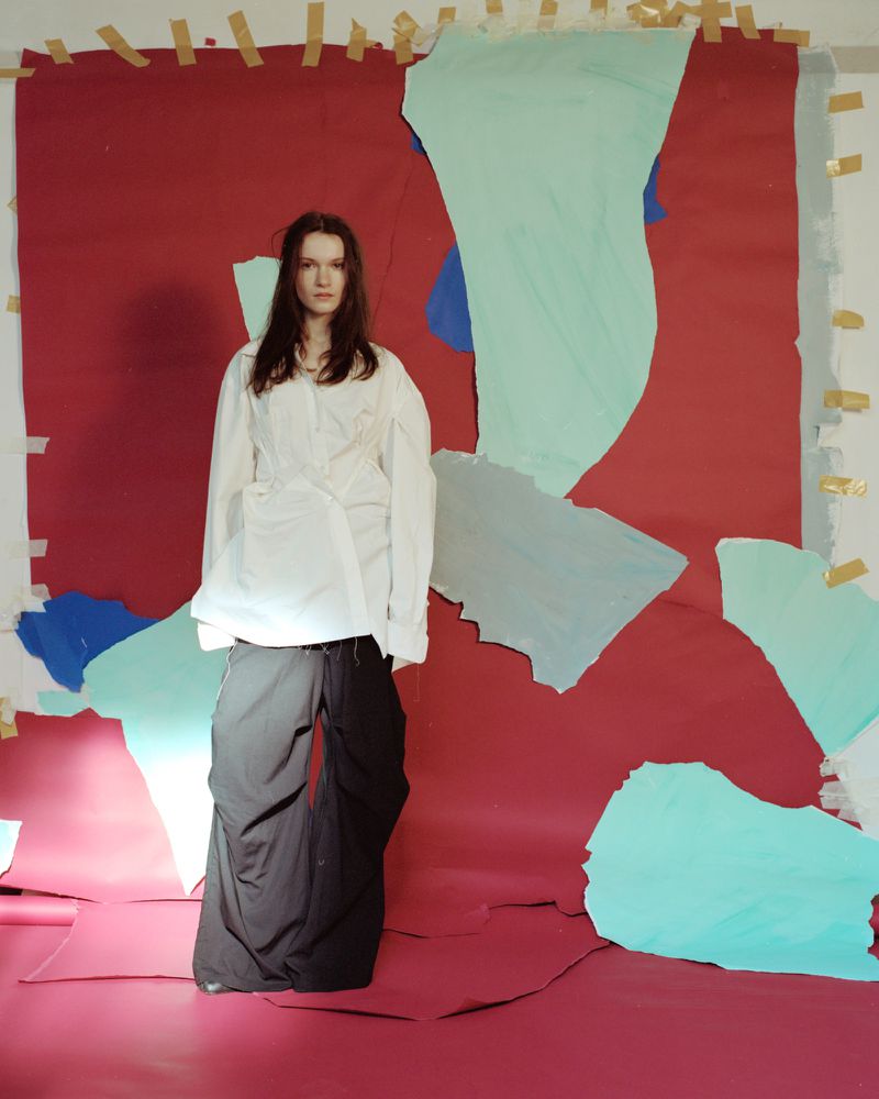 Fashion portrait of a young adult female in white shirt and black pants posing against a backdrop of colored paper pieces and masking tape.