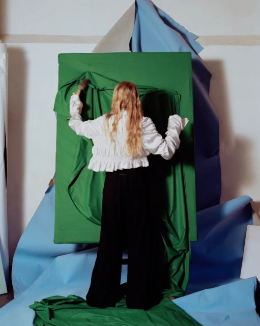 A woman faces away from the viewer, artfully arranging swathes of green and blue fabric against a white wall in an indoor setting.