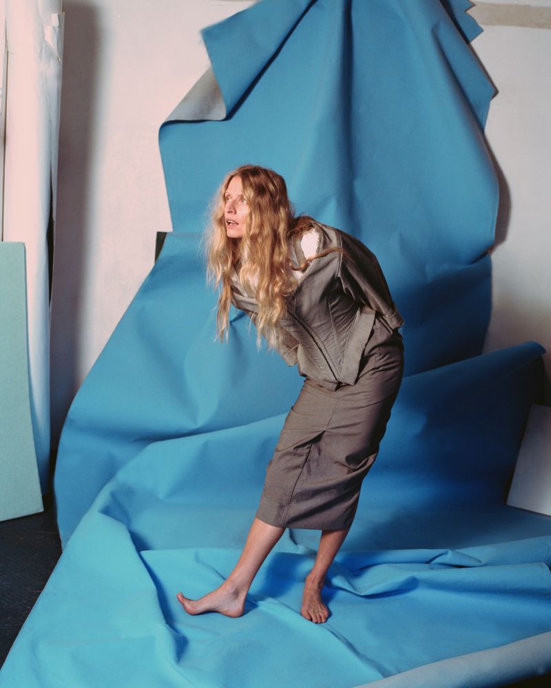Woman in a grey suit posing in front of a light blue backdrop. She is barefoot, wearing a corset-style top and skirt.