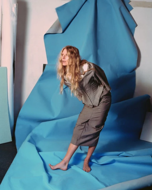 Woman in a grey suit posing in front of a light blue backdrop. She is barefoot, wearing a corset-style top and skirt.