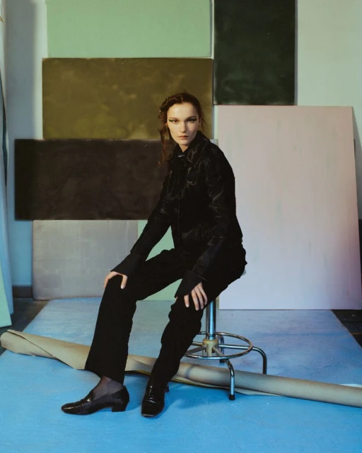 A woman in a dark outfit sits on a metal stool in a studio with neutral colored panels on the wall and a blue surface on the floor.