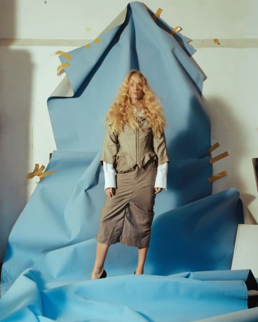 A fair-skinned, curly-haired woman in a beige skirt and jacket poses in front of a light blue fabric background. High-heeled shoes visible.