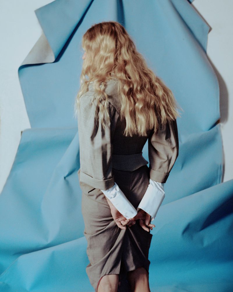 Back view of an adult woman in a grey skirt suit standing against a blue paper background in an indoor fashion shot.