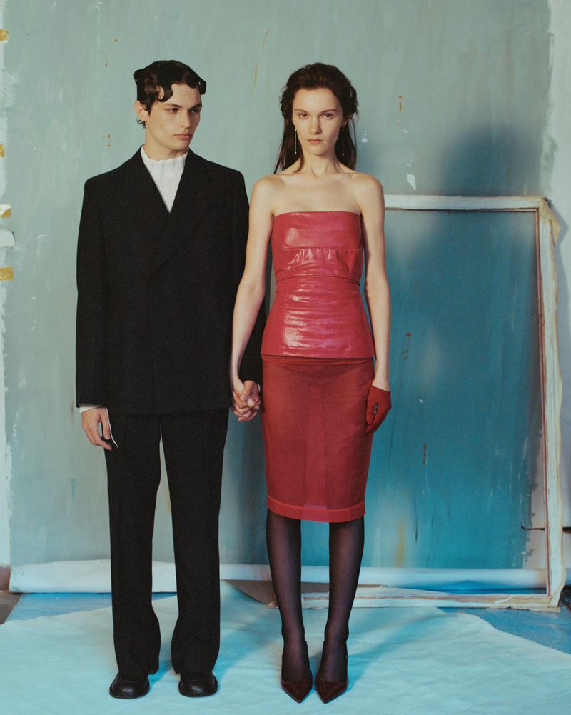 A man in a black suit and a woman in a red strapless dress holding hands against a painted blue wall