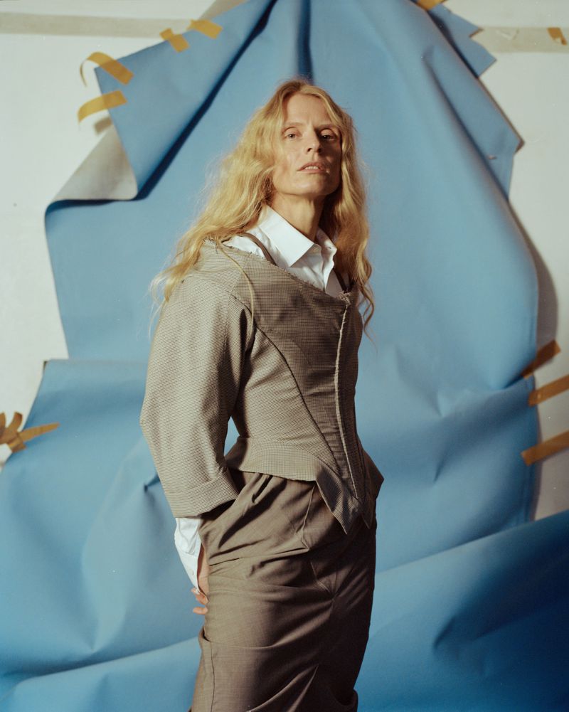 Portrait of a blonde-haired woman in a skirt suit with a collared shirt in front of a blue paper backdrop with yellow tape.