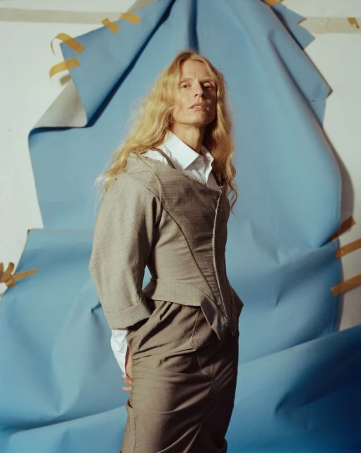 Portrait of a blonde-haired woman in a skirt suit with a collared shirt in front of a blue paper backdrop with yellow tape.