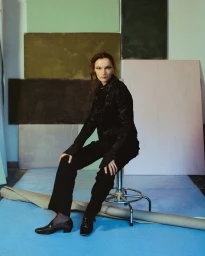 Model in black jacket and trousers sitting on a stool against a backdrop of pastel panels for Duel Magazine Scarlet Sails editorial.