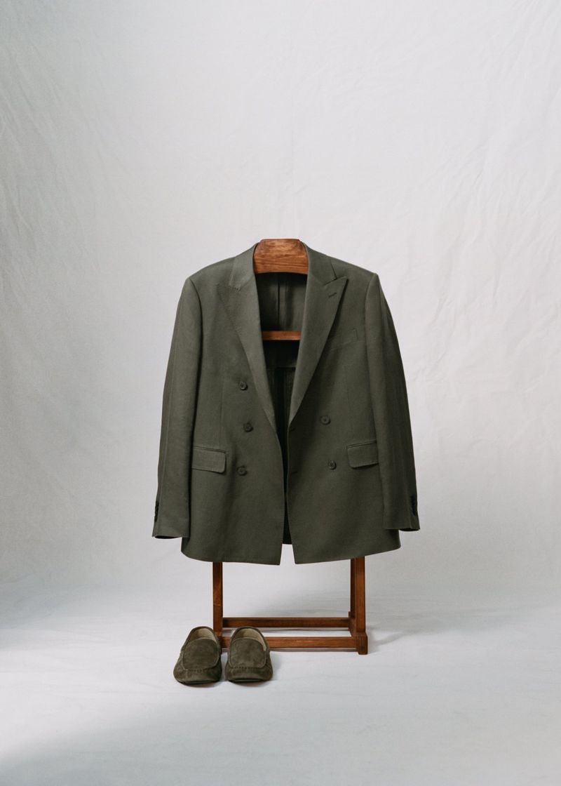 Olive green suit jacket on a wooden hanger, paired with suede loafers against a neutral backdrop in a softly lit indoor setting.