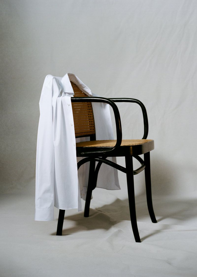 Minimalist shot of a black wood chair with a woven seat and a white shirt draped over the arm in front of a white backdrop.