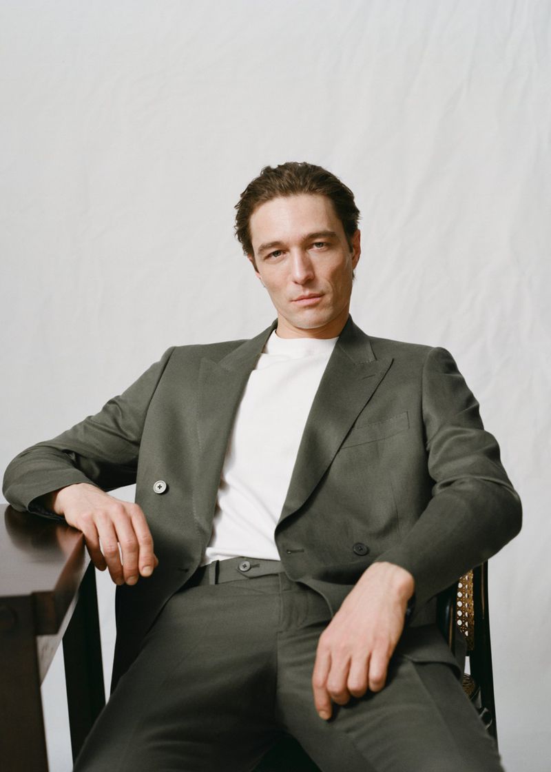 Adult male wearing a suit, seated indoors. The man is wearing a grey suit jacket and pants with a white T-shirt against a light background.