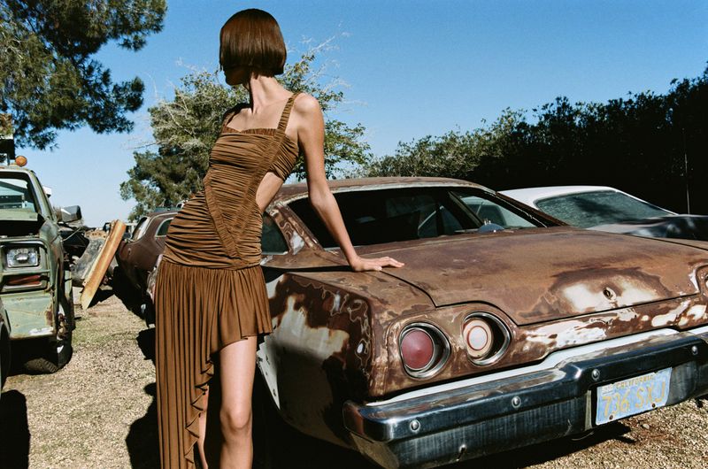Adult female model poses in a brown dress leaning on a rusty car in an outdoor junkyard setting on a sunny day with trees and blue sky.