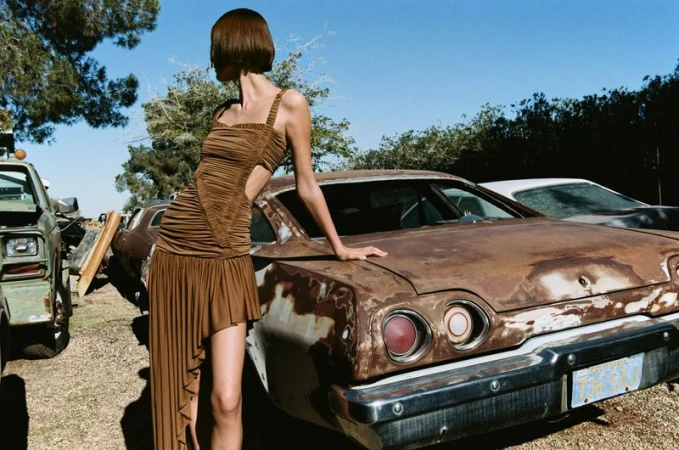 Adult female model poses in a brown dress leaning on a rusty car in an outdoor junkyard setting on a sunny day with trees and blue sky.
