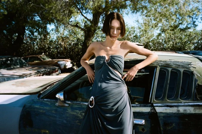 A young woman in a blue strapless dress poses against a worn car in an outdoor junkyard setting, under a bright midday sun with trees visible.