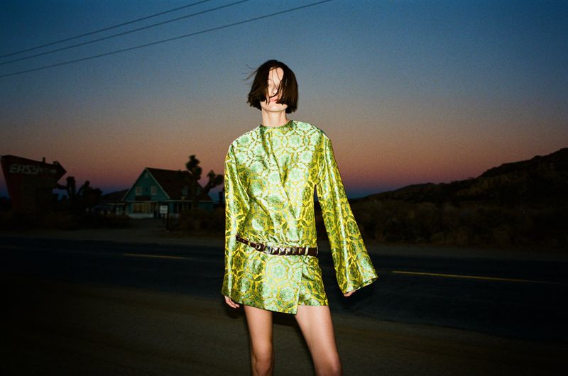 A woman in a green patterned mini dress with a belt stands on an empty road near a motel at dusk, fashion editorial photography.
