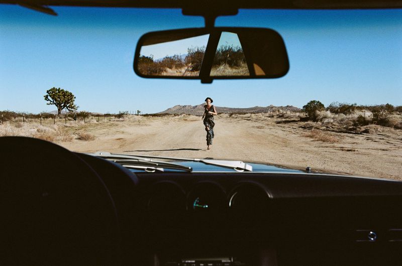 A woman in a dress is seen from inside a car, running barefoot on a dirt road through a desert landscape on a clear, sunny day.
