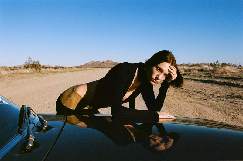 A brunette woman in black top rests on a vintage car against a desert backdrop with clear blue sky and distant hills. Calm and serene.