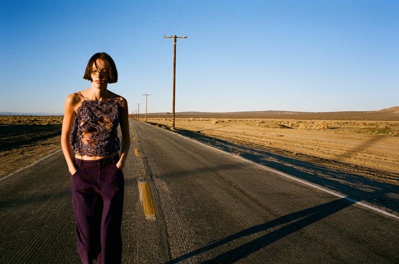 A slender female fashion model strides confidently on an empty desert road wearing a lace top and loose purple pants on a sunny day.