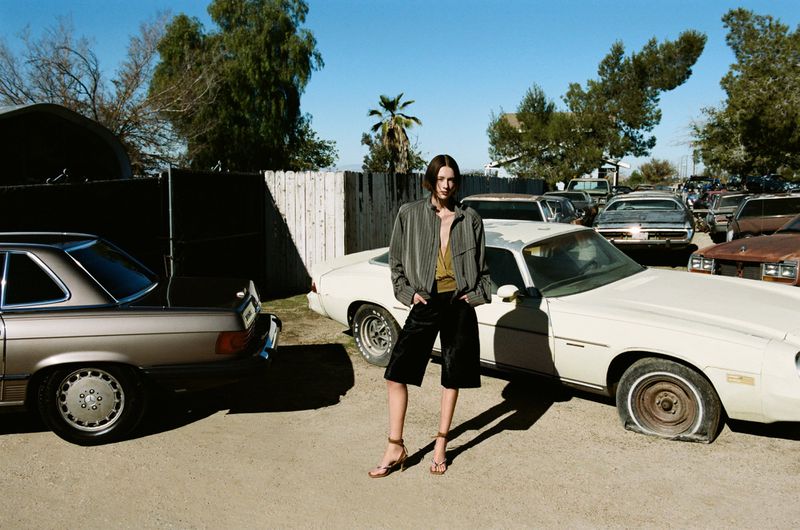 A woman posing in black shorts and a neutral top and jacket stands between two vintage cars in a daytime outdoor setting.