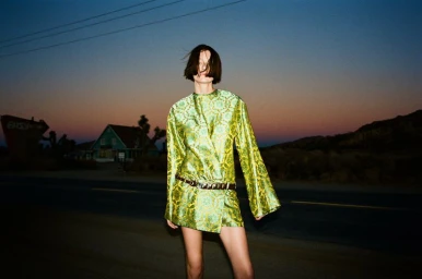 Model wearing a patterned green dress standing on a deserted highway during dusk with a colorful sunset sky.