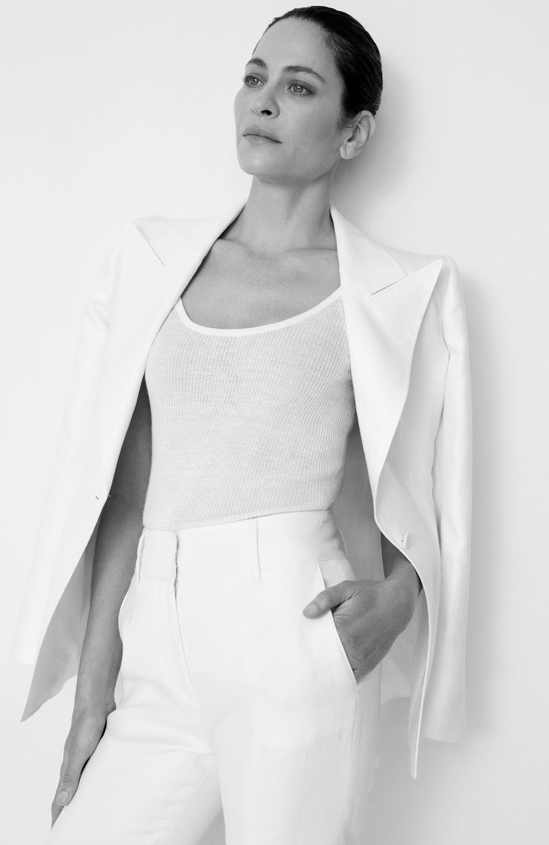 Black and white portrait of an adult woman in a white suit with hand in pocket against a white background in eye-level shot.