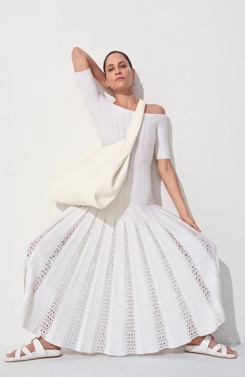 Woman poses in white knit dress and matching sandals with white shoulder bag, against a white backdrop in a fashion shot.