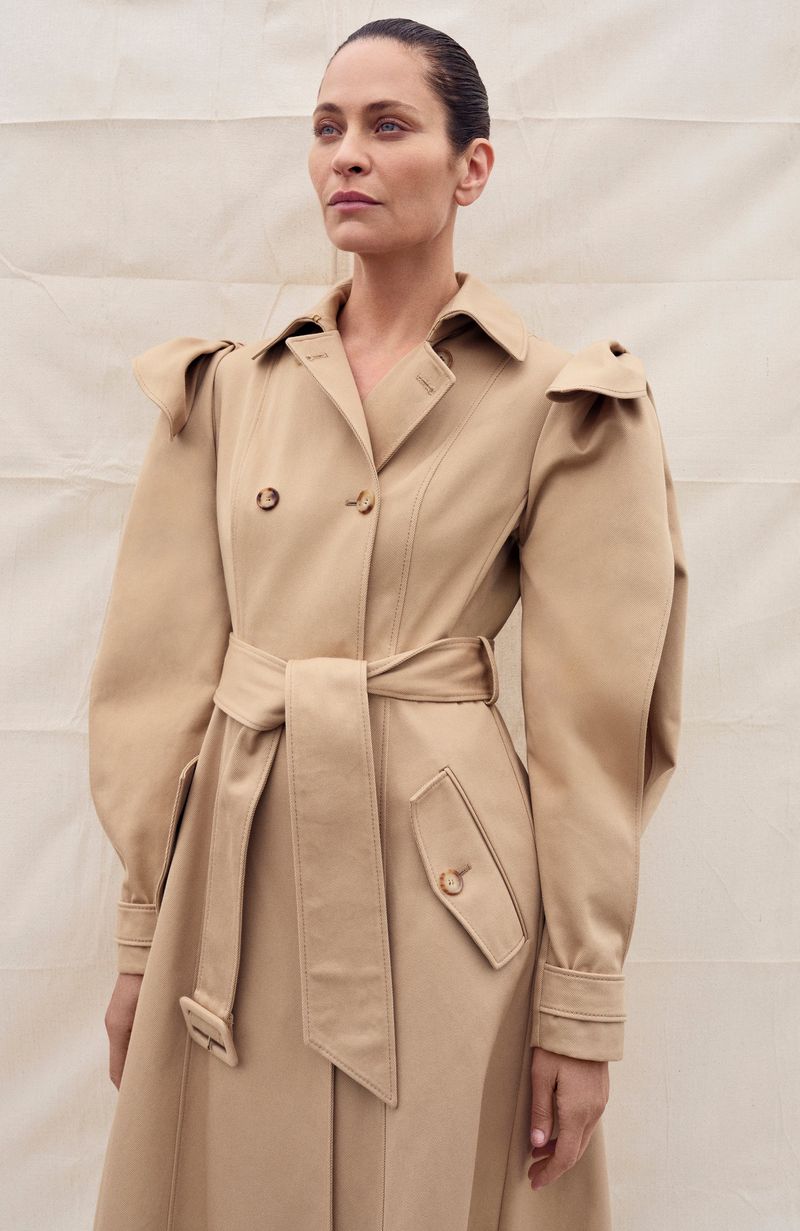 Adult woman wearing a tan belted trench coat poses in front of an off-white textured background in a medium shot.