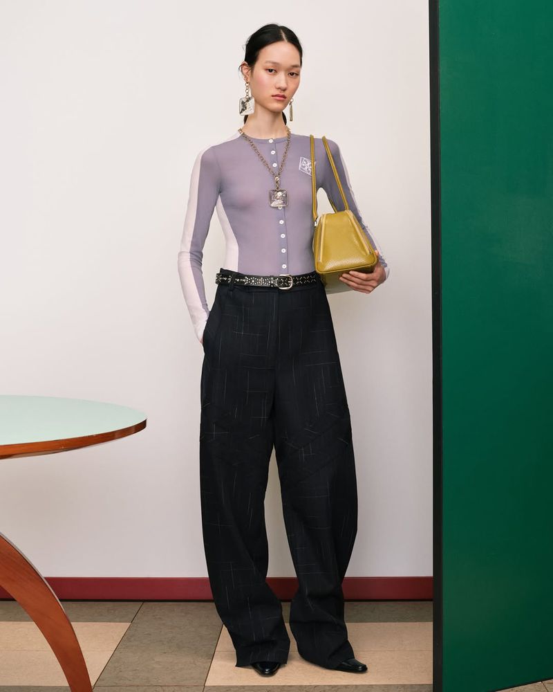 Fashionable woman in wide-leg pants, cardigan, and statement jewelry holds a yellow purse against a neutral studio backdrop.