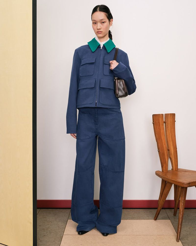 A woman in blue jacket and pants with a brown purse stands in a studio setting with a chair on a neutral-colored surface.