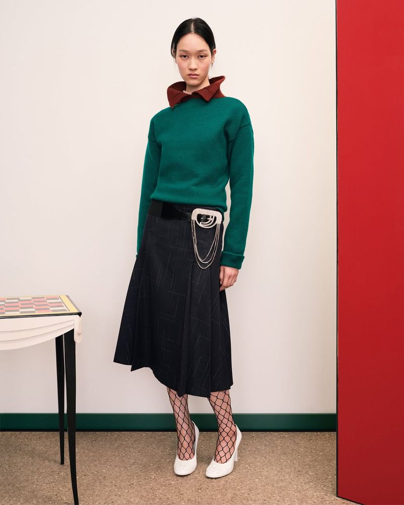 Adult female fashion model poses indoors wearing a green sweater with brown collar, black skirt, fishnet stockings, and white high heels.