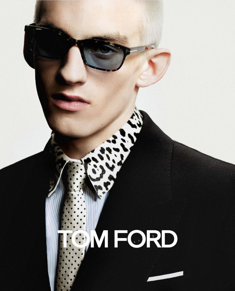 Close-up portrait of a blond male model wearing a patterned shirt, tie, black blazer, and dark sunglasses, featuring TOM FORD text.