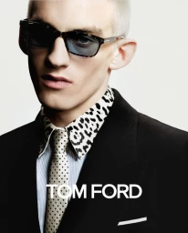 Close-up portrait of a male model wearing tortoiseshell sunglasses, a black jacket, patterned shirt, and tie against a white background.