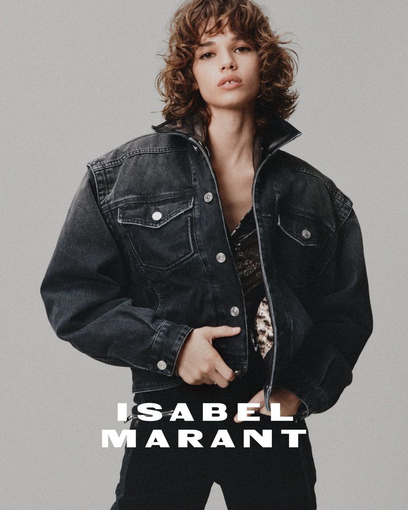 Fashion shot of a woman with curly brown hair wearing a denim jacket and black pants in front of a neutral studio backdrop.