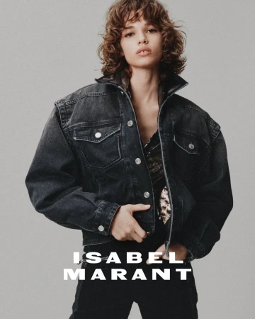Fashion shot of a woman with curly brown hair wearing a denim jacket and black pants in front of a neutral studio backdrop.