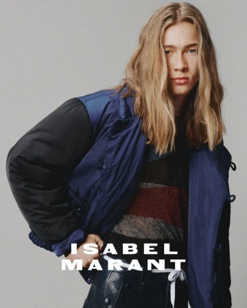 A young man with shoulder-length blonde hair wears a navy and black bomber jacket over a striped sweater against a plain gray background.
