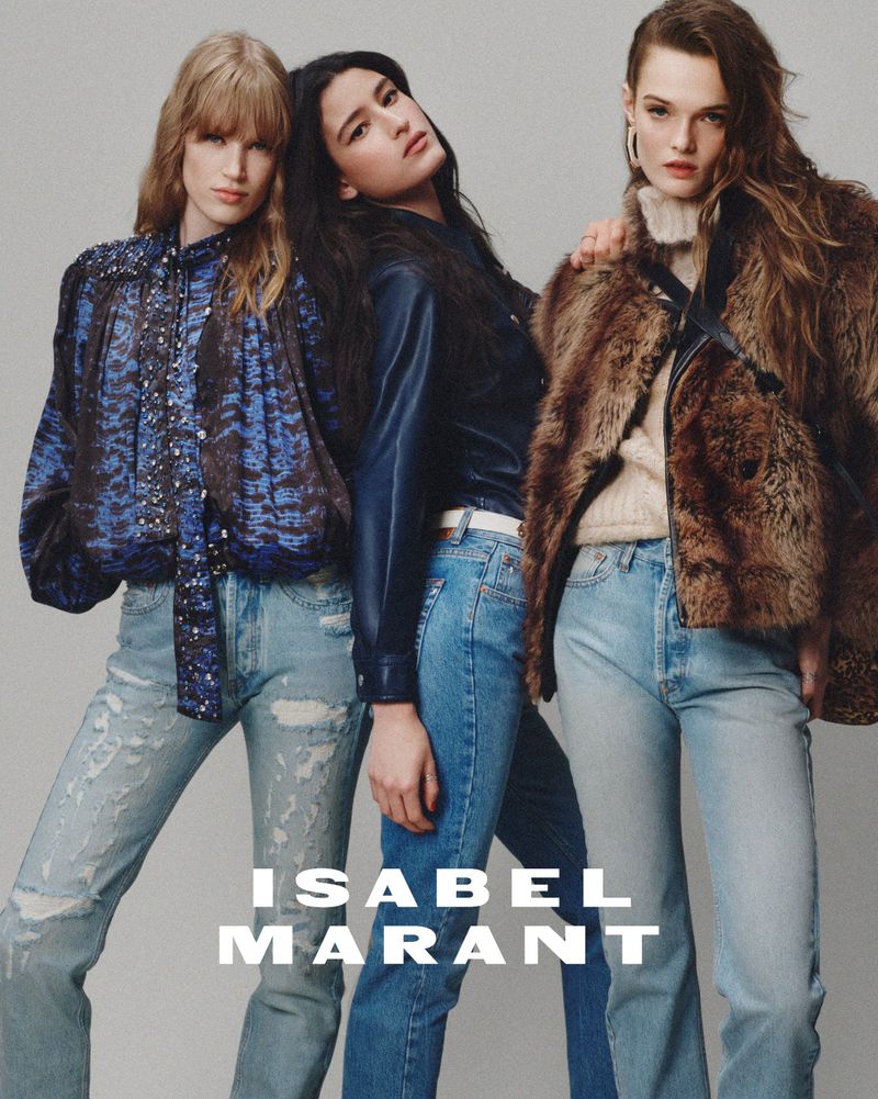 Three fashion models pose together in light-wash jeans and jackets in a studio shot for Isabel Marant, showcasing contemporary apparel.